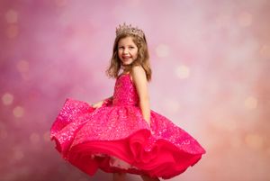 Young child twirls in sparkly hot pink pageant dress and tiara against pink bokeh background.
