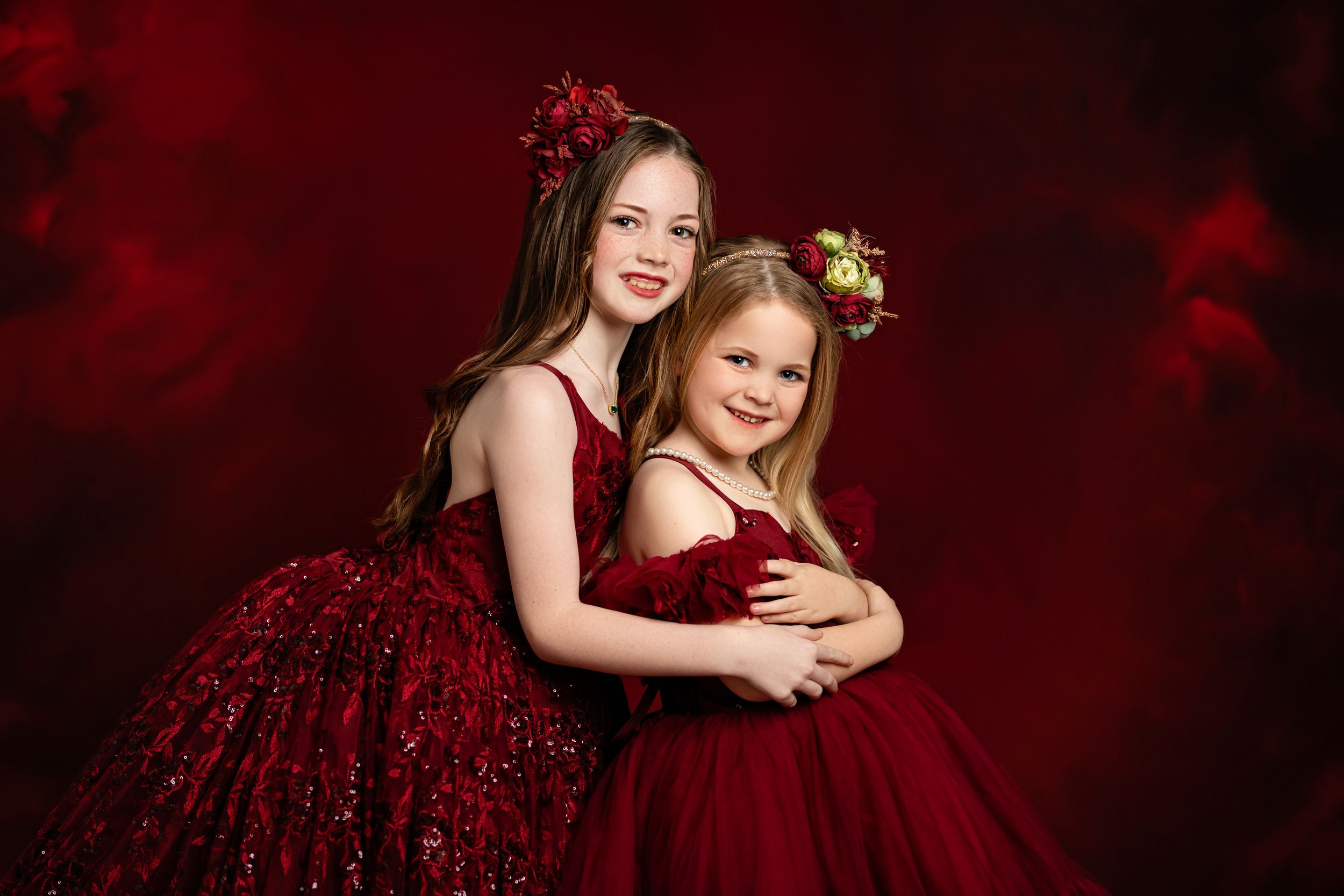 Two people in elegant burgundy dresses pose together against a dark red backdrop in a glamorous portrait setting.