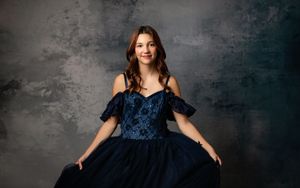 A person poses in an elegant navy blue gown with off-shoulder design against a textured gray backdrop.