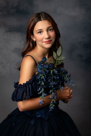 Studio portrait of someone in an elegant navy blue off-shoulder gown holding blue wisteria flowers against a dark background.