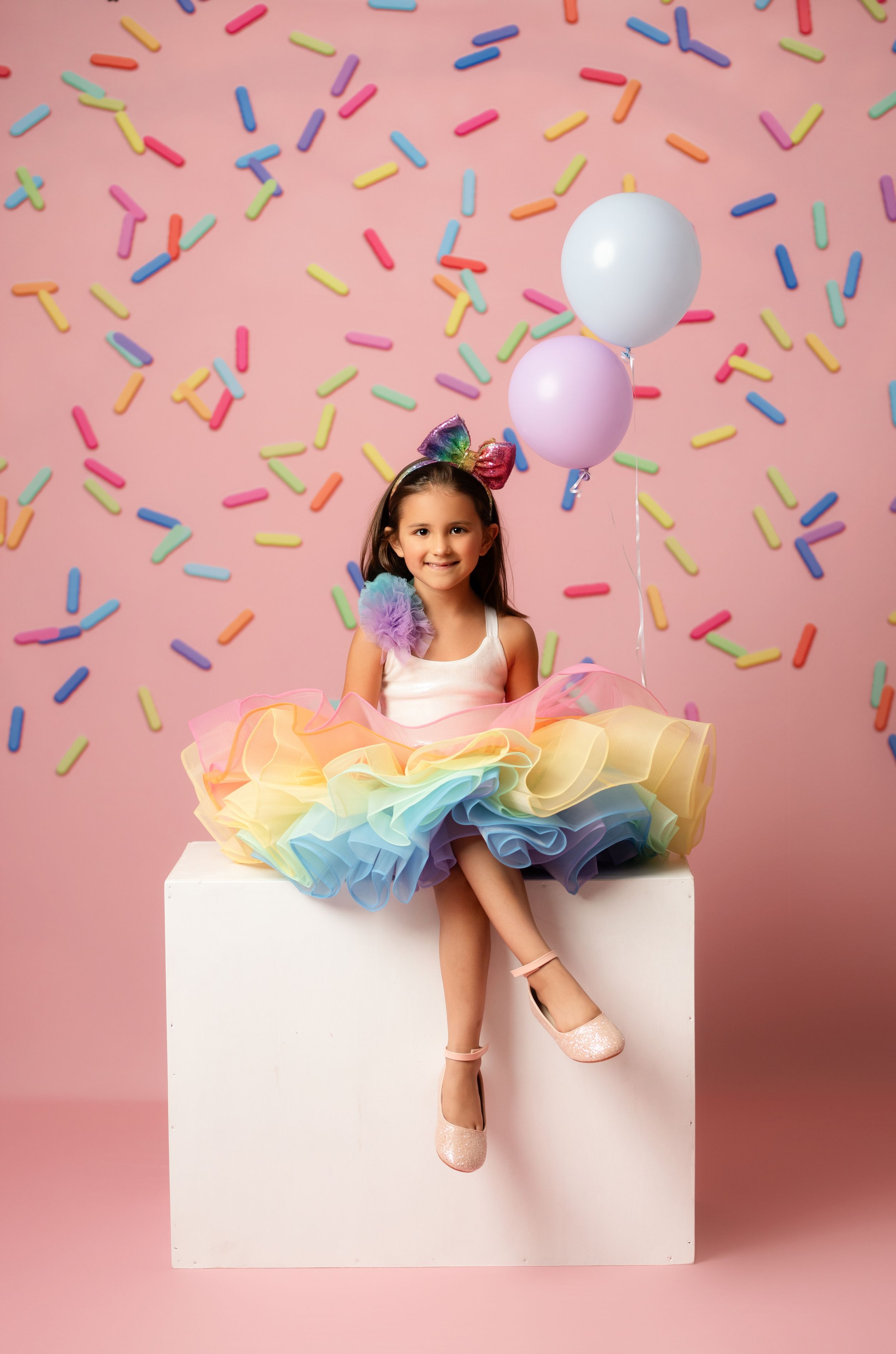 A smiling girl in a rainbow tutu dress sits on a white cube, holding balloons against a pink sprinkles backdrop.