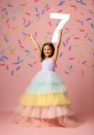 A girl in a rainbow tulle gown holds a number 7 sign with colorful sprinkles falling on a pink background.