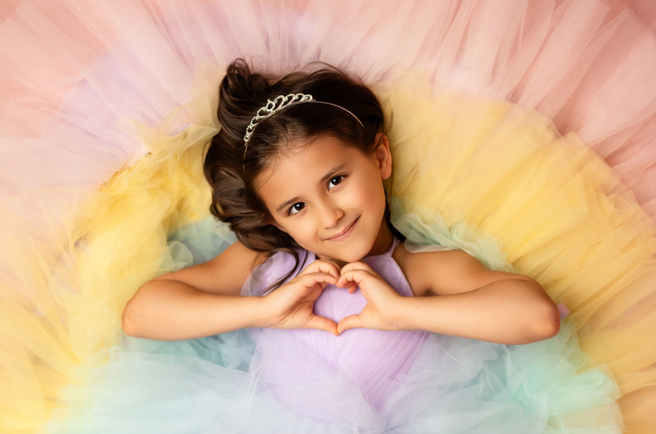 Young girl in colorful rainbow tutu dress making heart shape with hands, wearing tiara, smiling at camera.