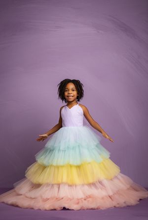 A young child twirls in a flowing rainbow pastel tulle dress against a purple studio backdrop.
