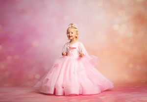 A young girl in a pink princess gown and tiara smiles joyfully against a soft bokeh pink background.