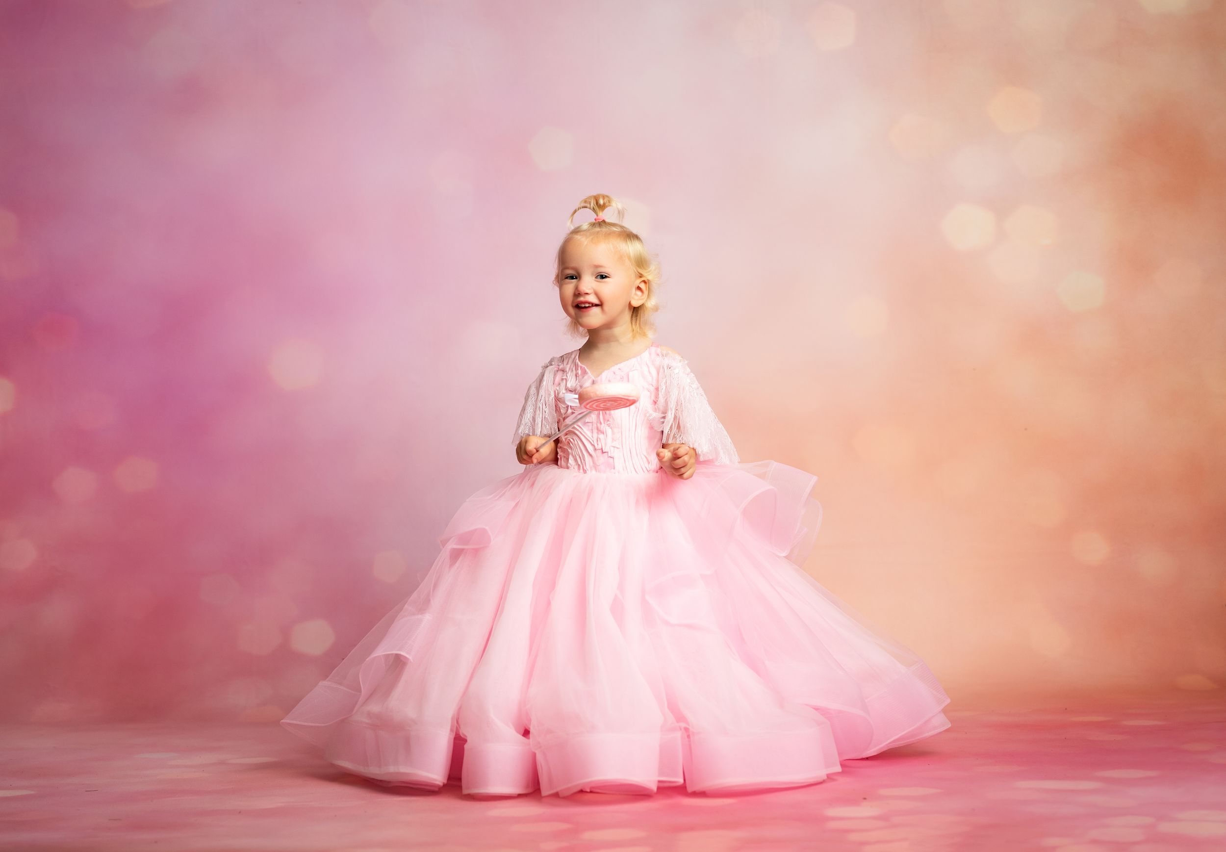 A young girl in a pink princess gown and tiara smiles joyfully against a soft bokeh pink background.