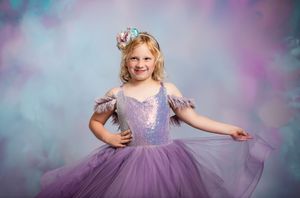 Young dancer in a sparkly purple tulle dress poses gracefully against a dreamy pastel background.