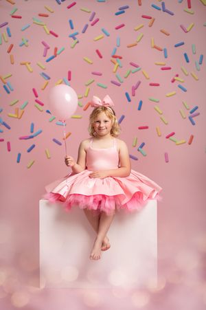 Young child in pink tutu dress smiles against backdrop with colorful confetti sprinkle pattern.