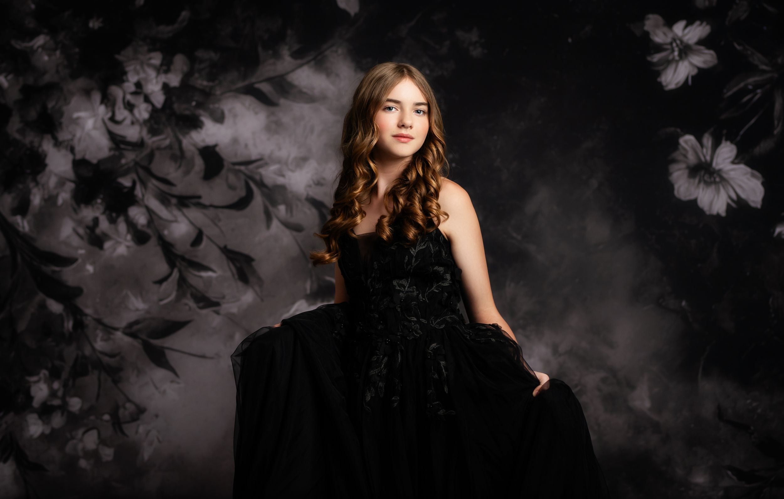 Young woman in elegant black gown poses gracefully against a dark floral backdrop in a professional portrait.