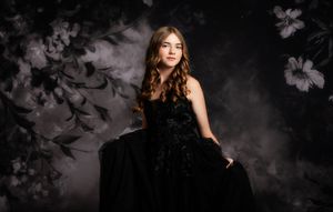 Young woman in elegant black gown poses gracefully against a dark floral backdrop in a professional portrait.