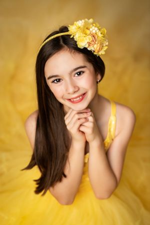 Portrait against yellow background wearing a yellow dress and floral hairpiece with a gentle smile.