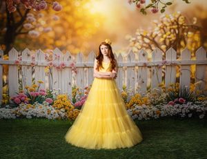 A yellow tulle ballgown dress stands out against a garden backdrop with white picket fence and blooming flowers at sunset.