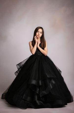 A model poses in an elegant black tulle ball gown with layered ruffles against a neutral studio backdrop.