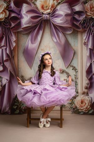 A dreamy photo setting with large purple satin bows and roses featuring someone in a lavender ruffled dress seated on a stool.
