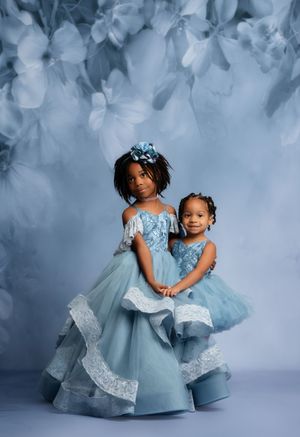 Two young girls in matching blue tulle gowns pose elegantly against a soft floral blue backdrop.