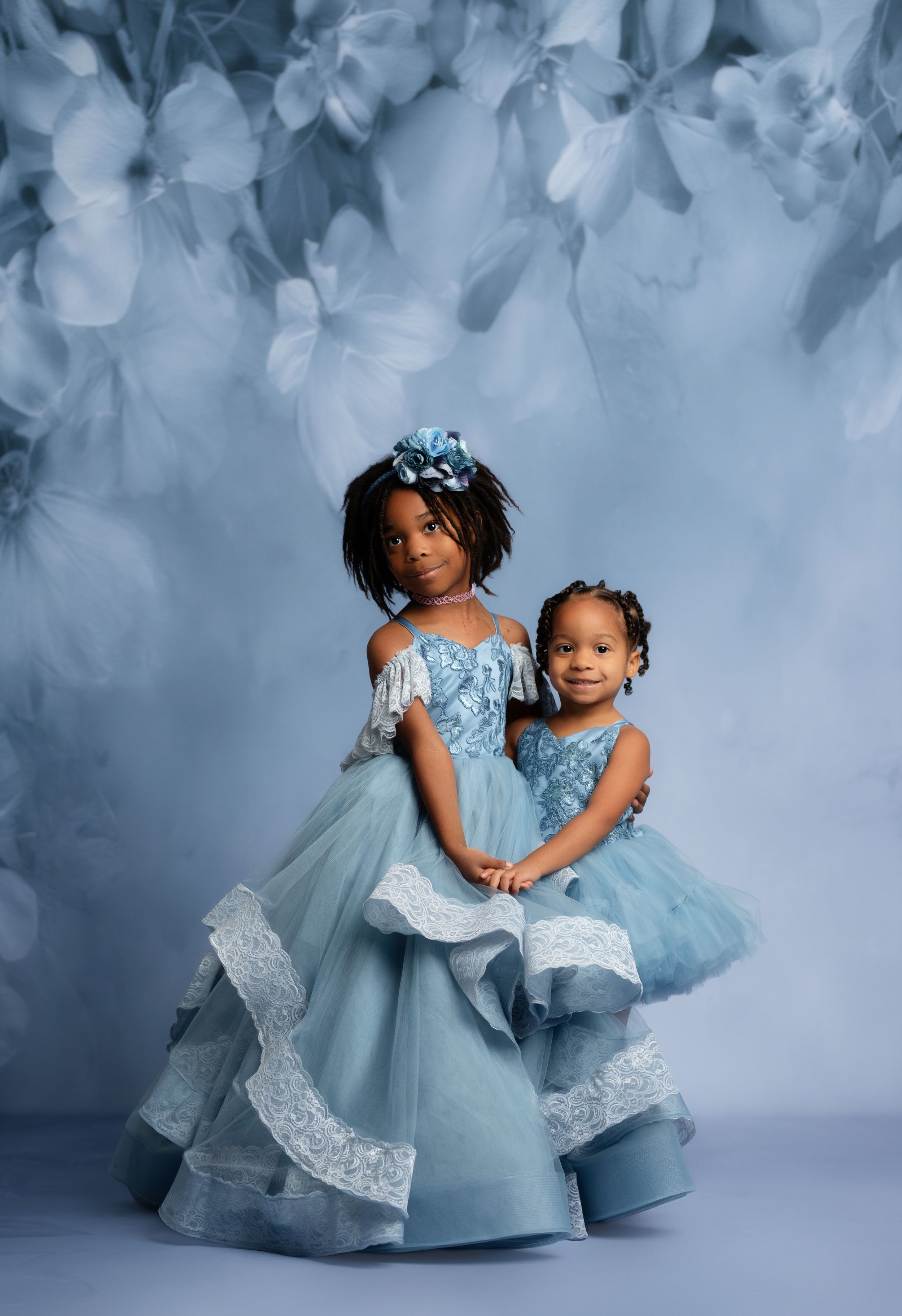 Two young girls in matching blue tulle gowns pose elegantly against a soft floral blue backdrop.
