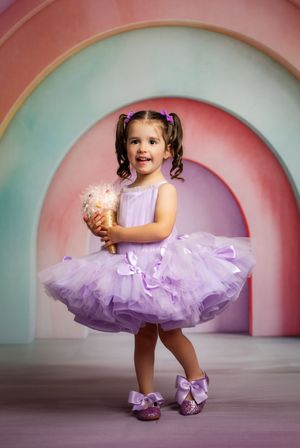 Young girl in purple tutu dress holding ice cream cone, posing before colorful rainbow arch backdrop.