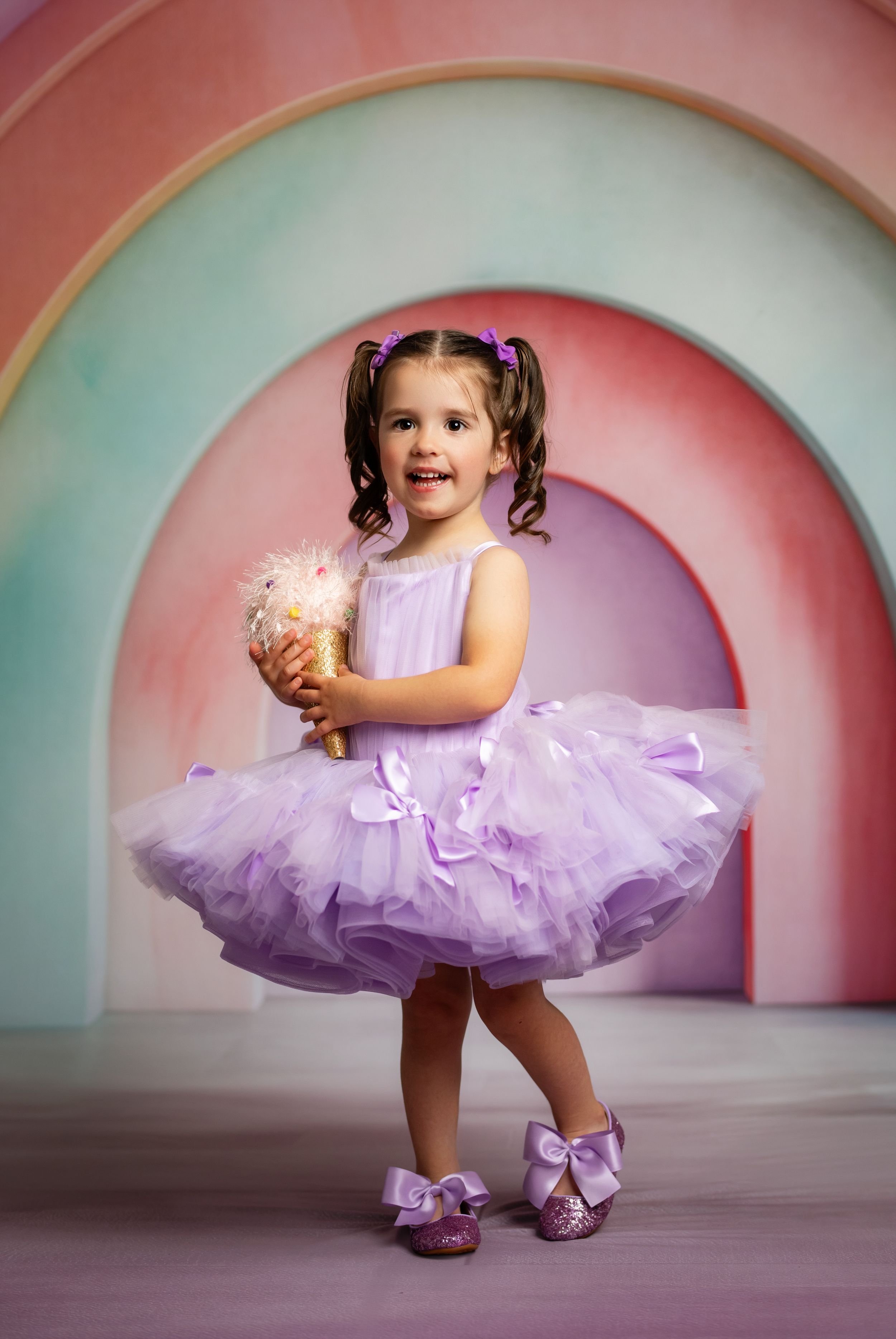 Young girl in purple tutu dress holding ice cream cone, posing before colorful rainbow arch backdrop.
