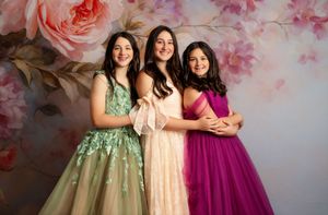 Three young women in elegant ball gowns pose together in front of a floral backdrop.
