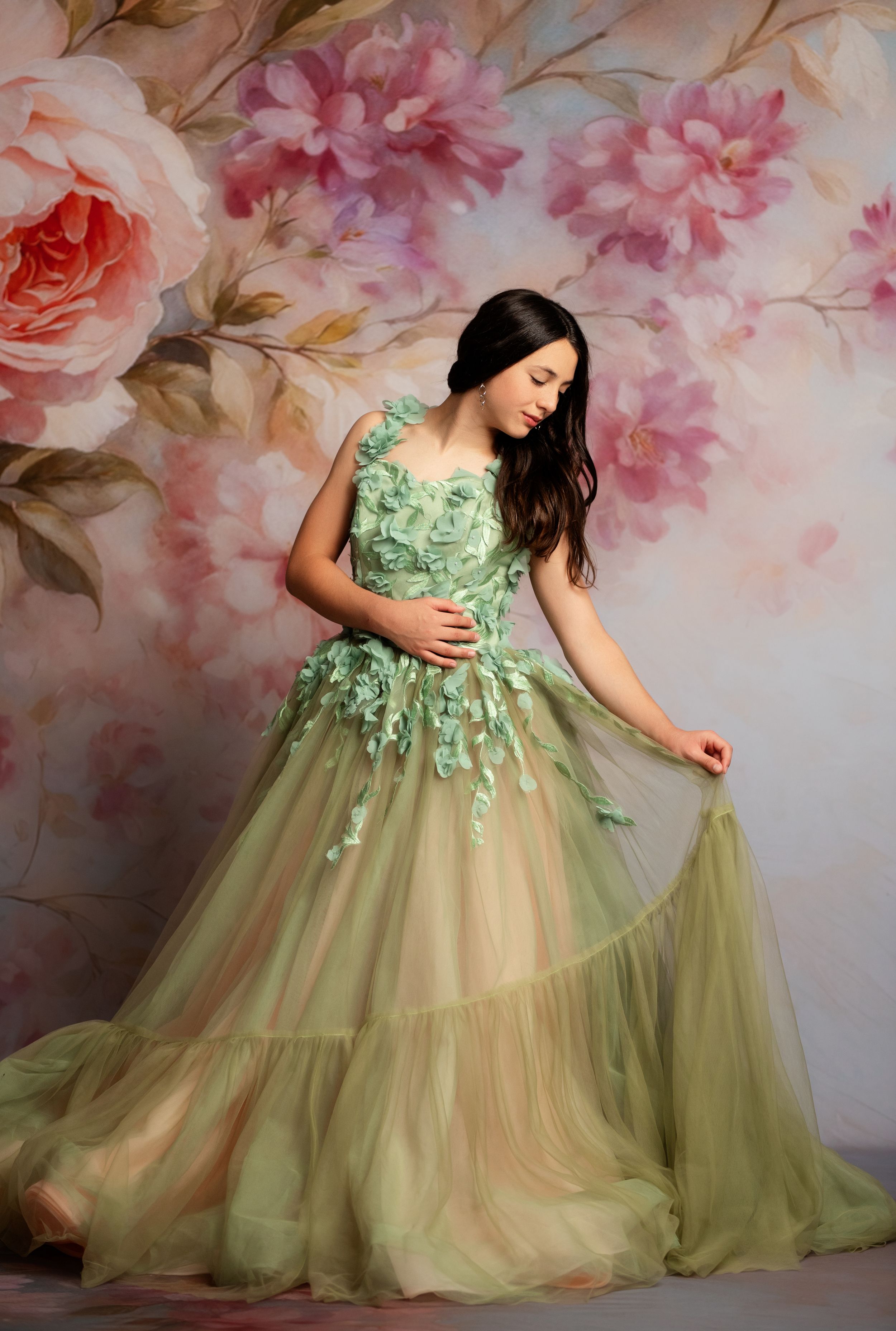 Young woman in elegant green floral tulle gown poses gracefully before a soft pink rose floral backdrop.