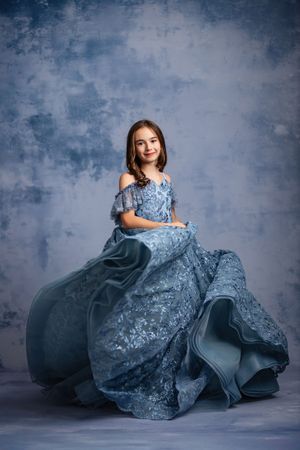 A elegant blue lace gown with off-shoulder design billows dramatically against a textured grey backdrop.