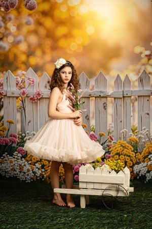Dreamy garden scene with tulle dress, white picket fence and flowers at sunset with golden bokeh lighting effect.