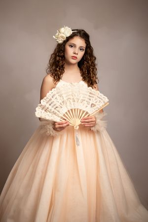 A figure in a flowing peach-colored tulle gown poses elegantly with a decorative lace fan against a neutral backdrop.