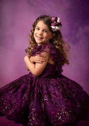 Young model wearing a sparkly purple ball gown with floral hair accessory against a purple studio backdrop.
