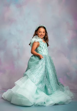 A child wearing a stunning mint green ball gown with lace details and ruffled tulle layers poses against a dreamy background.