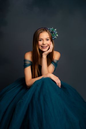 Smiling girl in teal off-shoulder ball gown with floral hair accessory and braces, posing gracefully in studio.