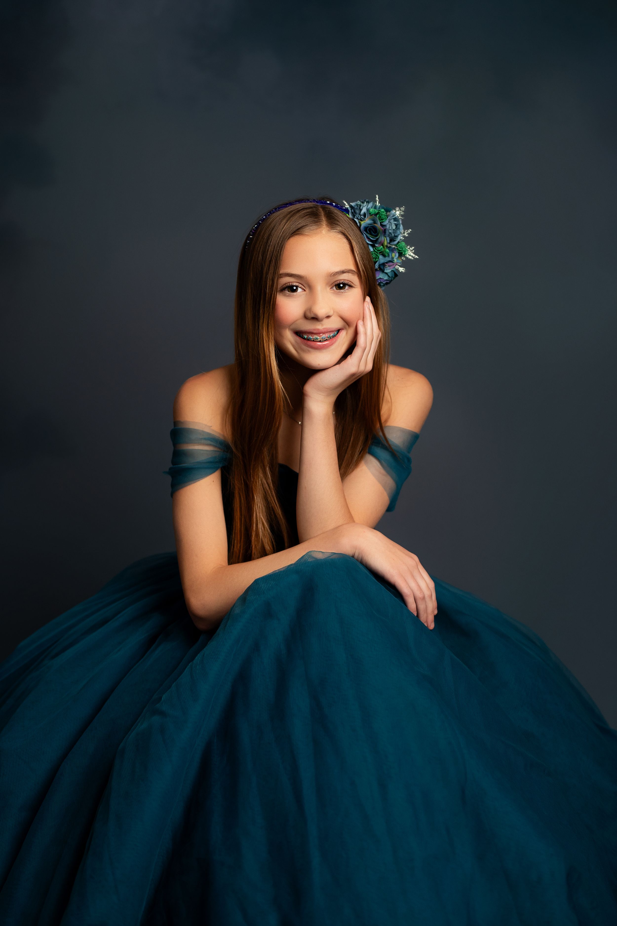 Smiling girl in teal off-shoulder ball gown with floral hair accessory and braces, posing gracefully in studio.