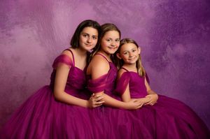 Three girls in matching magenta tulle ball gowns pose together against a purple backdrop in a professional portrait.