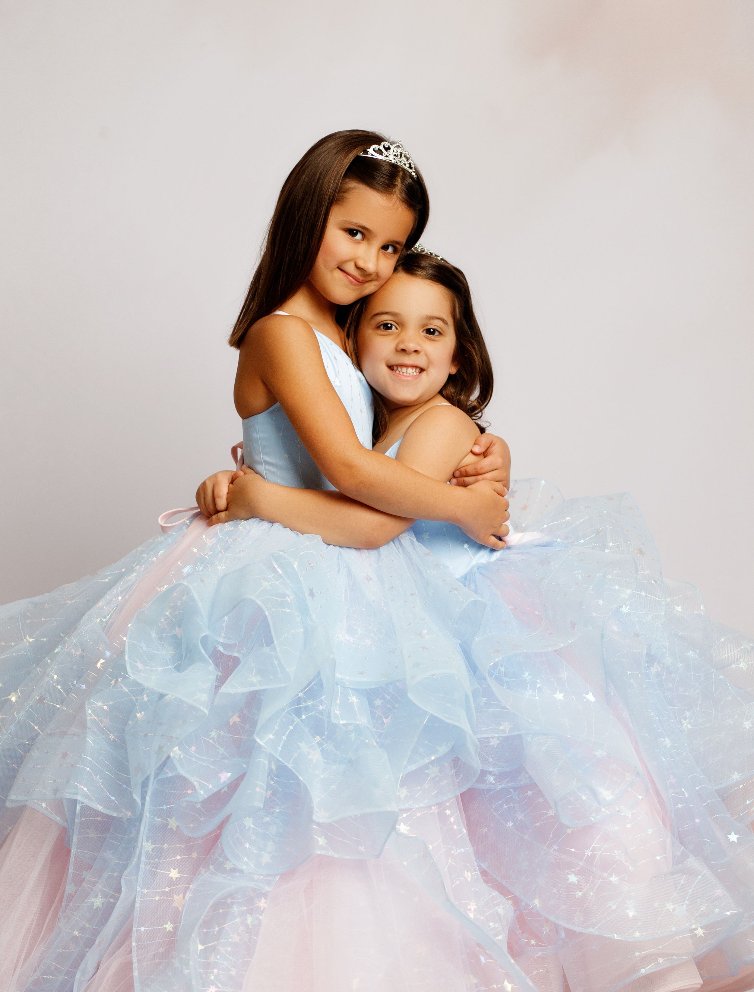 Two young girls in sparkly blue layered ball gowns embrace, one wearing a tiara, in a studio portrait.