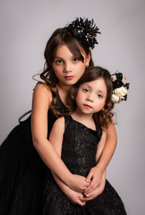 Two children in matching black formal dresses adorned with floral headpieces pose together for a portrait.