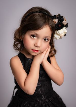Studio portrait of a young child in an elegant black dress with flowers in their hair against a gray backdrop.