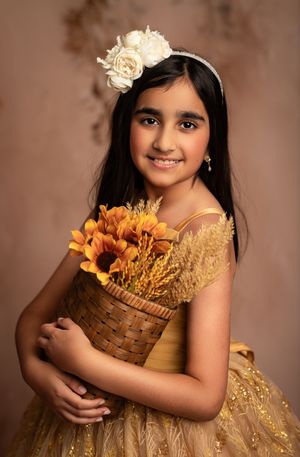 A child in a golden dress holds a woven basket filled with autumn flowers against a warm brown backdrop.