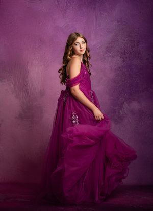 A young child twirls in a flowing plum-colored dress against a purple textured backdrop in a series of graceful poses.