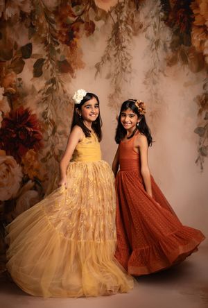 Two children in contrasting yellow and rust-colored dresses twirl and pose together against a floral backdrop.