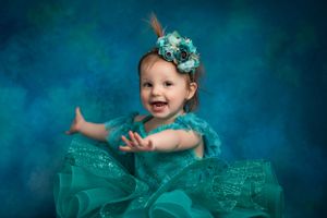 A smiling baby wearing a turquoise tulle dress and floral headpiece poses against a deep blue studio backdrop.