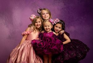 Four young children in formal dresses with ruffles pose together against a purple backdrop with pink floral accessories.
