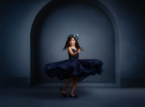 A ballerina in a flowing navy blue dress twirls gracefully in an illuminated stone archway against a dark background.