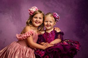 Two adorable children in pink outfits and flower headbands laugh and embrace while posing against a purple backdrop.
