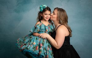 A touching moment captured in a studio portrait featuring a black dress and turquoise floral dress against a blue backdrop.