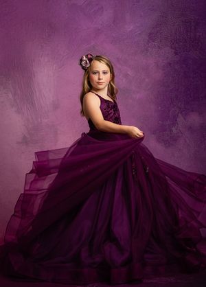 A dramatic portrait in purple tones featuring an elegant full-length gown with flowing tulle skirt against a textured background.