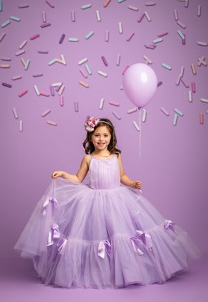 A purple princess dress twirls playfully as colorful sprinkles fall against a lavender backdrop in a dreamy portrait.