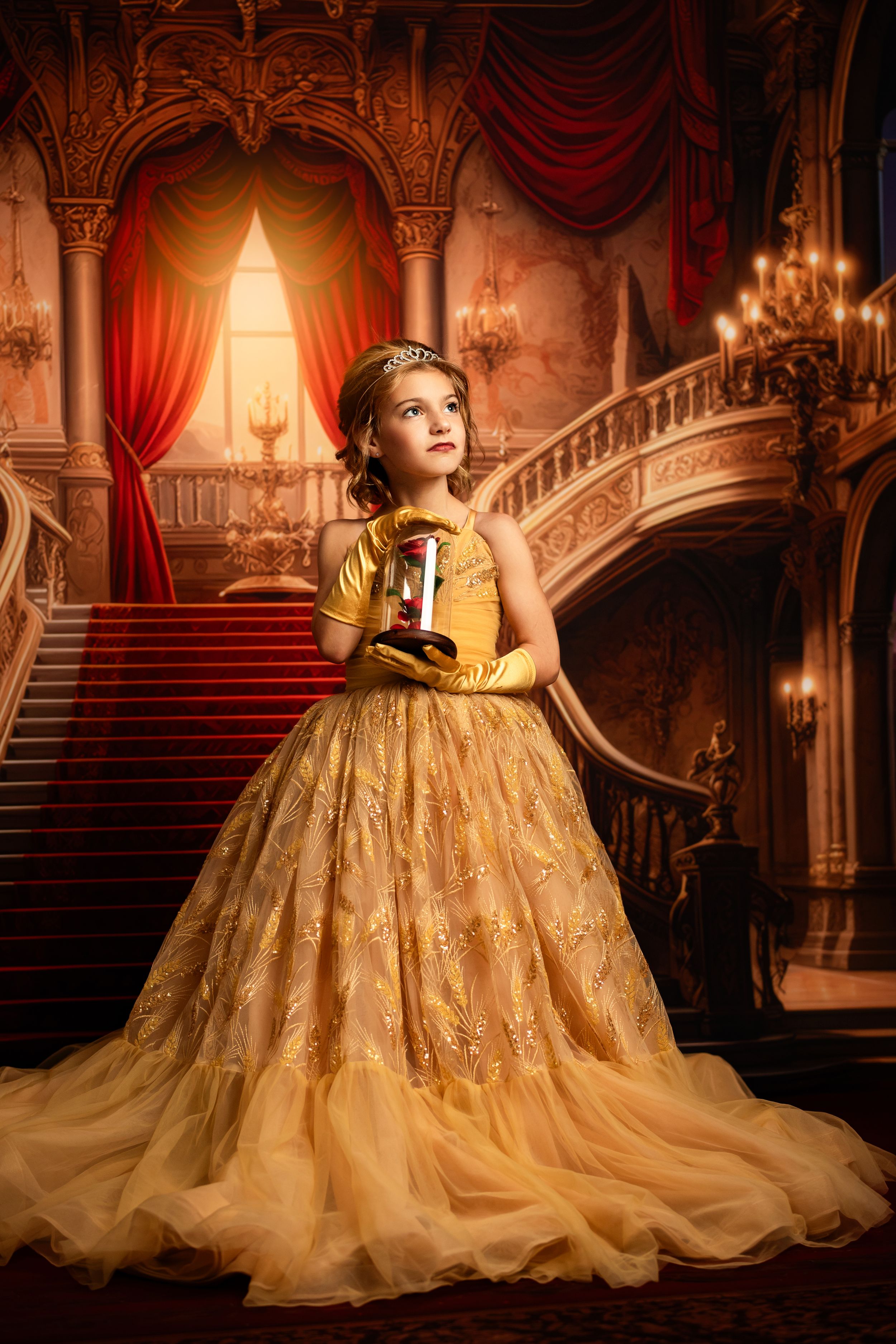A young girl in a golden Belle princess gown holds a lantern in an ornate ballroom with grand staircases.
