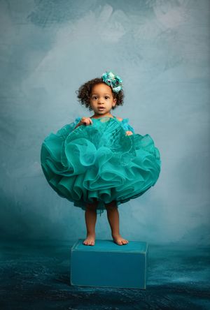 Small child wearing a turquoise ruffled dress and matching hair bow stands on a blue platform against a teal backdrop.