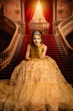Young girl in golden Belle princess gown and tiara poses regally on grand staircase with red carpet and ornate decor.