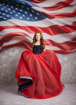 A patriotic portrait in front of an American flag backdrop featuring a flowing red and navy formal gown.