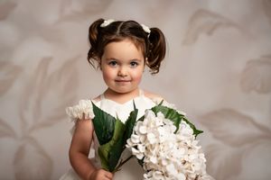 A child in a white dress poses with a large bouquet of white hydrangea flowers against a beige backdrop.